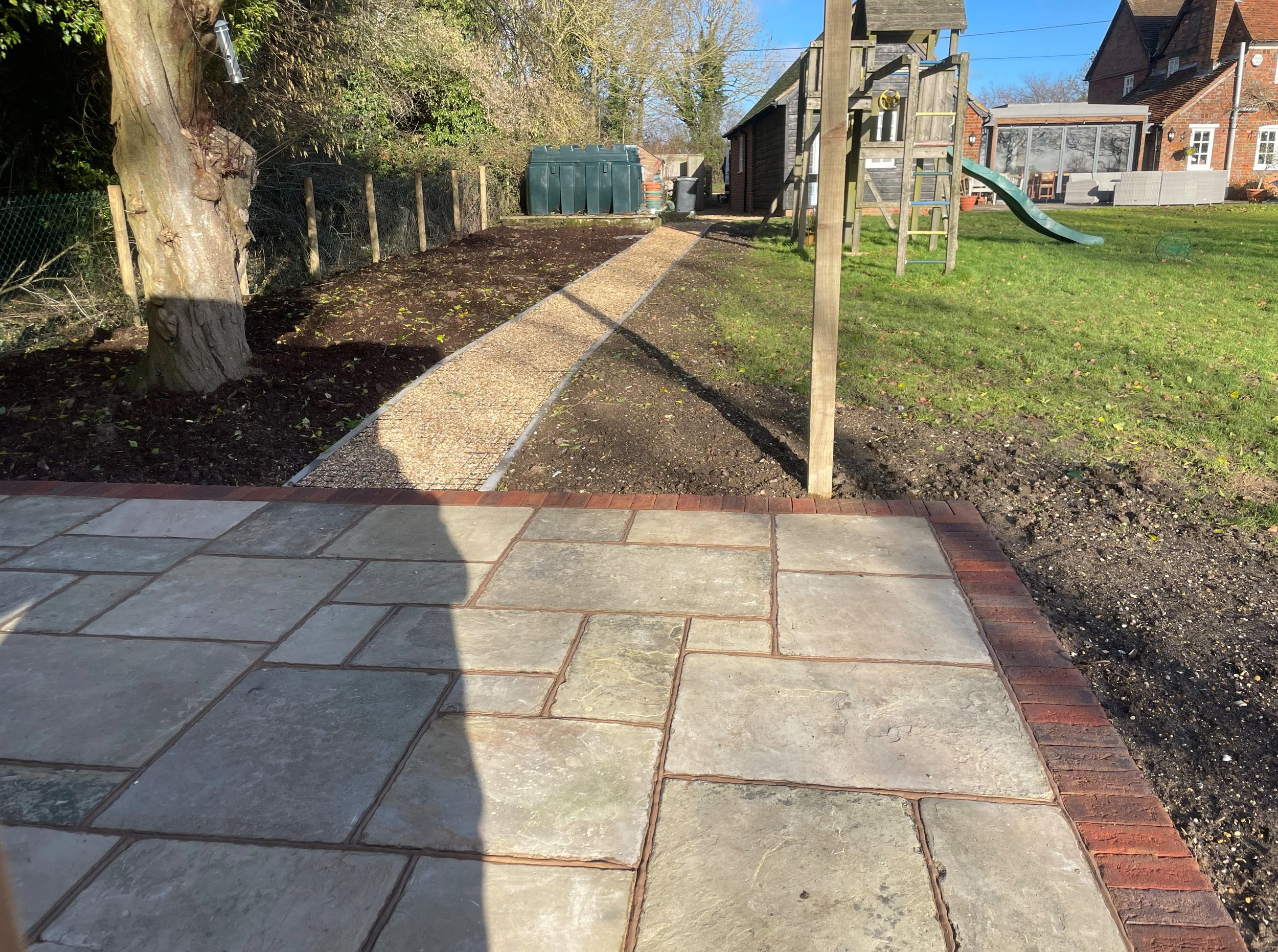 The image showcases a landscaped garden with a stone patio, gravel path, and green grass. Sunlight enhances the serene atmosphere
