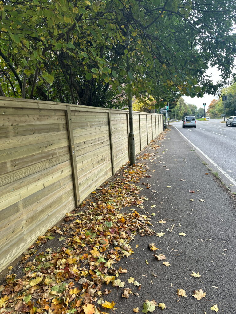 Wooden fence lining a sidewalk, surrounded by colorful autumn leaves and a road with passing cars