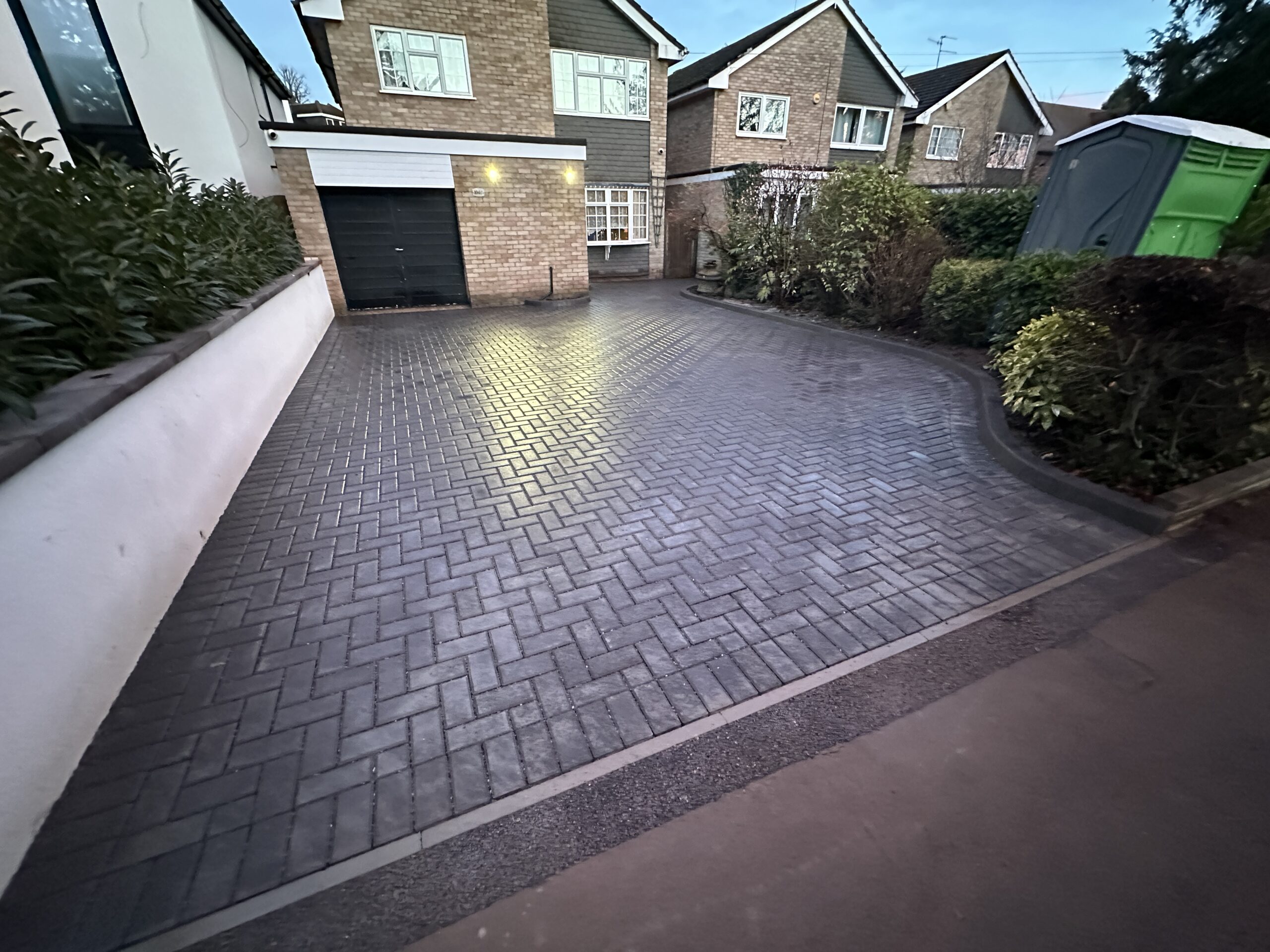 Spacious driveway with herringbone pattern paving, bordered by lush greenery and modern residential architecture, perfect for parking