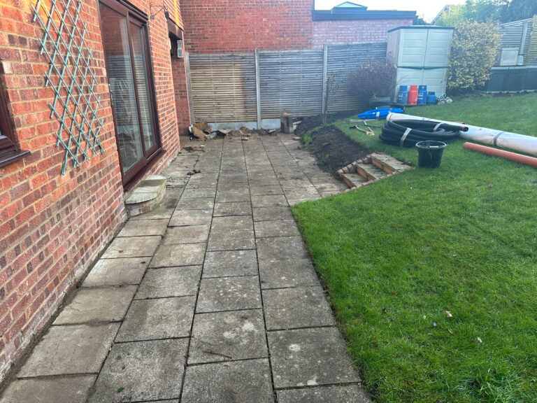 The image shows a garden area with a paved patio beside a house, featuring scattered tools and soil