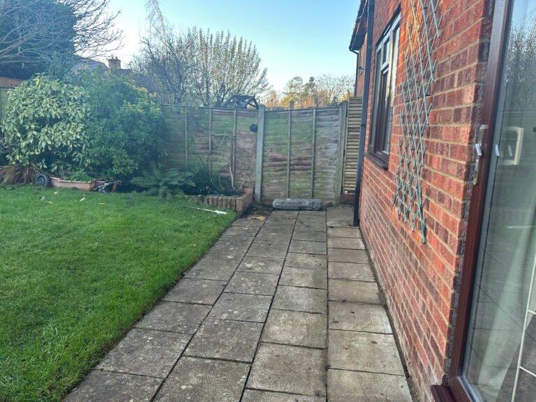 A serene garden pathway lined with paving stones, featuring lush greenery, bushes, and a wooden fence in the background