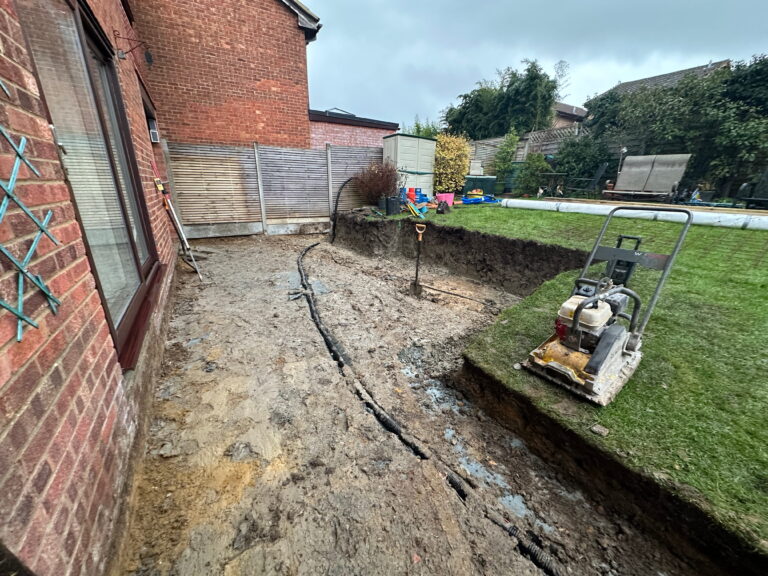 A partially excavated backyard features tools and machinery, with grass and garden elements visible in the background