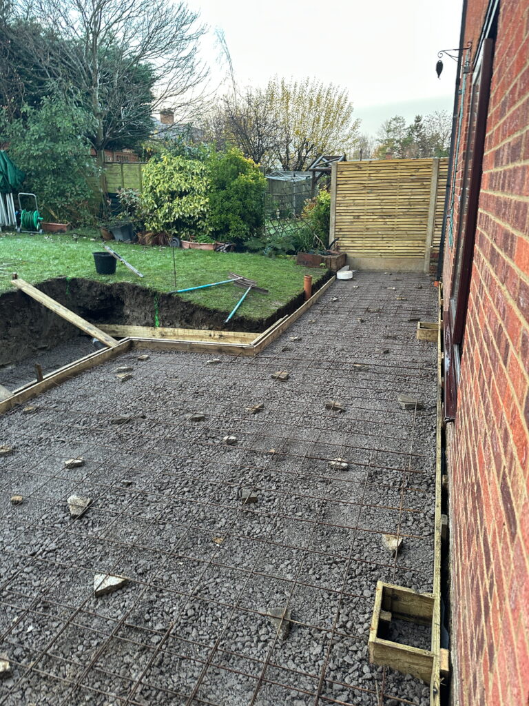 A construction site features a gravel foundation with rebar, tools scattered, and a grassy garden in the background