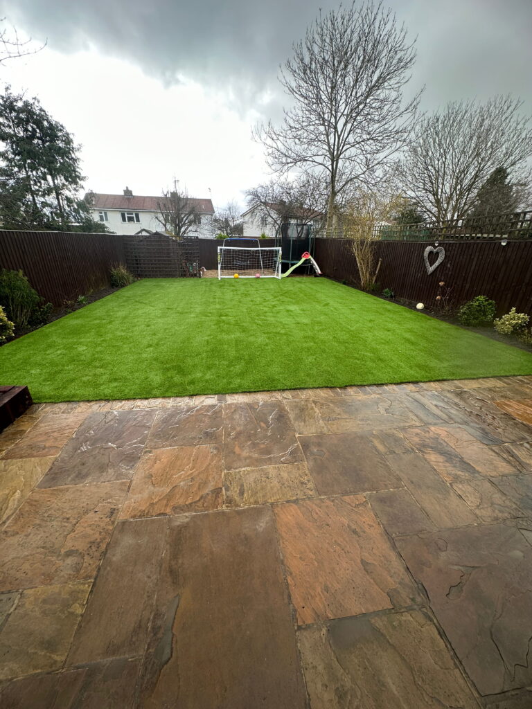 Lush green artificial grass garden featuring a soccer goal, kids’ toys, and a rustic stone patio under a cloudy sky
