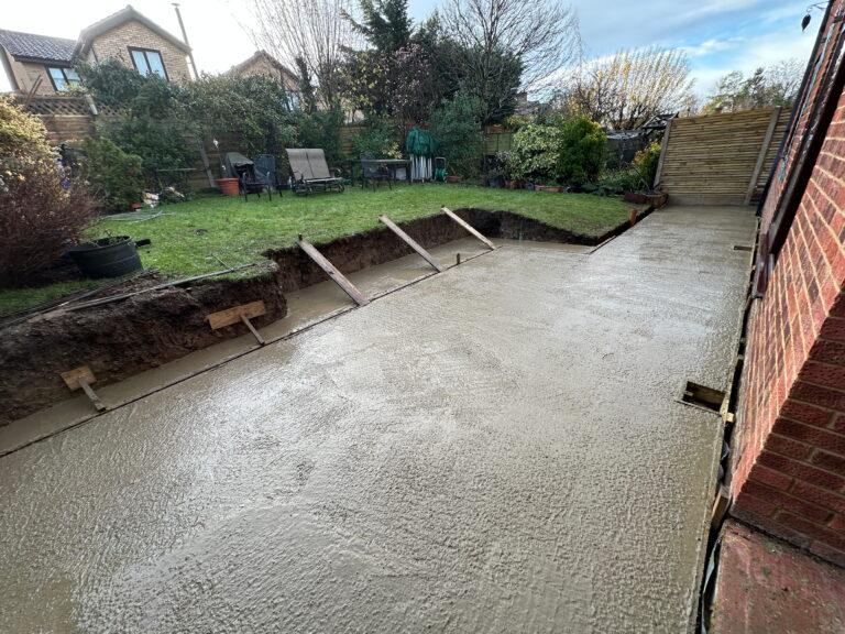 The image shows a freshly poured concrete slab in a backyard, with wooden framing and a grassy area in the background