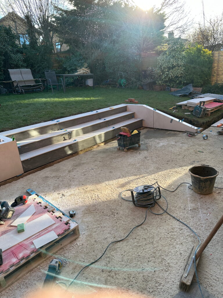 A partially constructed outdoor space with stairs, tools scattered around, and a lush garden backdrop. Sunlight is shining