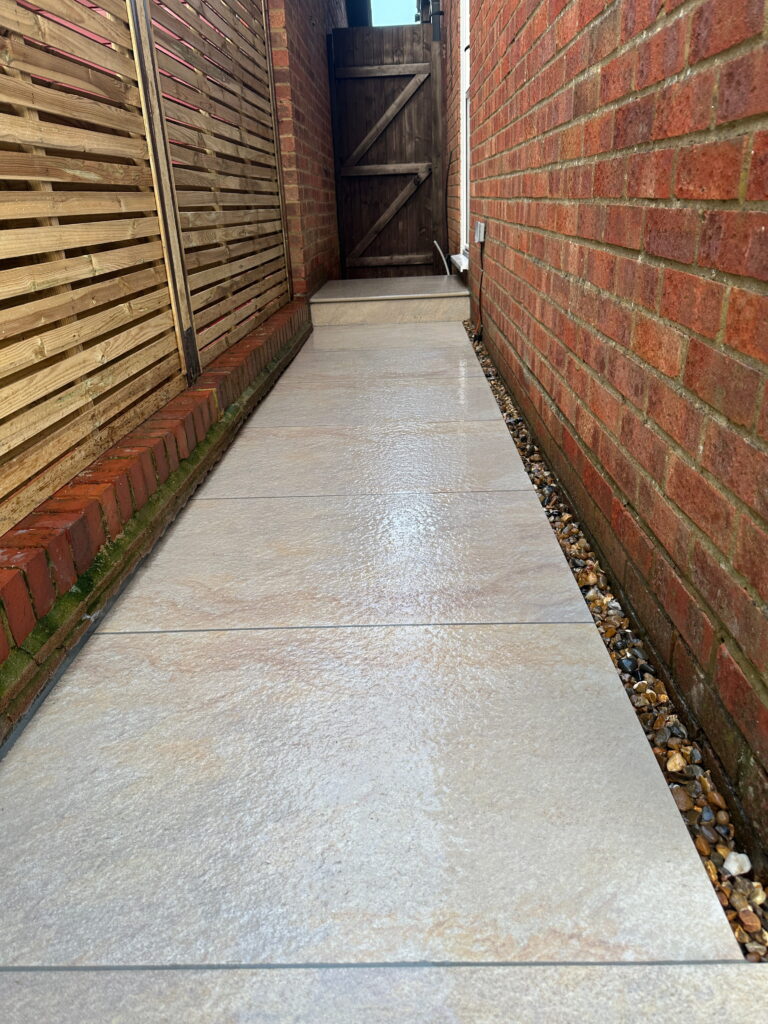 A narrow, paved pathway leads to a wooden gate, bordered by red brick walls and wooden fencing