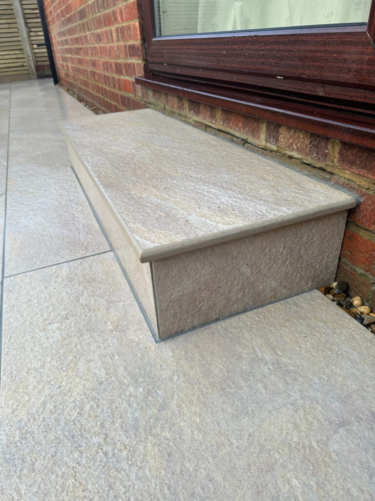 A smooth, beige stone step sits against a brick wall, framed by tiled flooring and a window above
