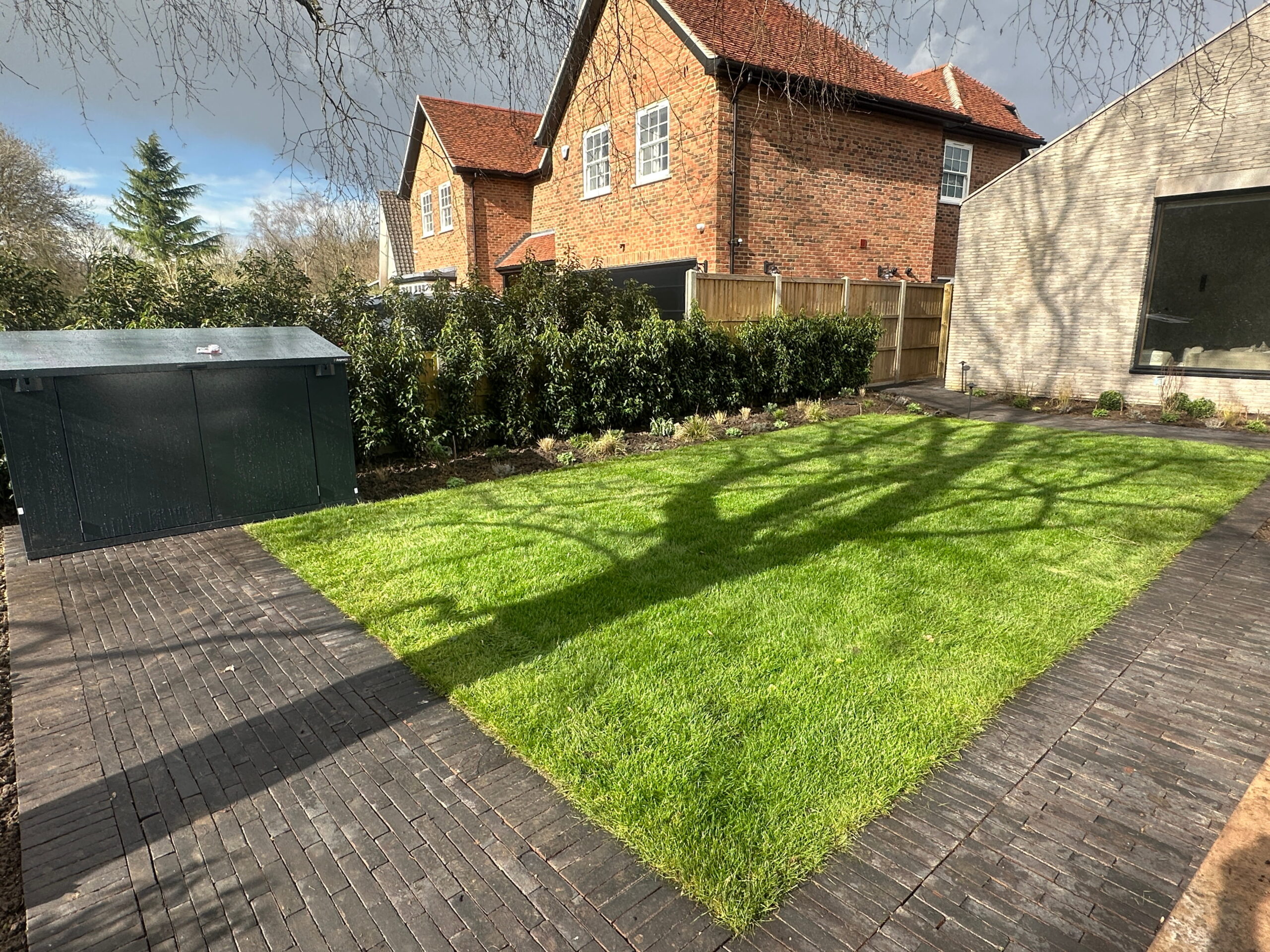 A well-maintained garden featuring lush green grass, brick pathways, and a storage shed, bordered by plants and two houses