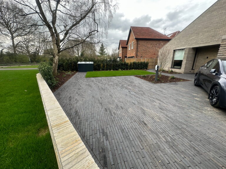 A modern driveway features a gray paver surface, green lawn, landscaping, and an electric car charging station