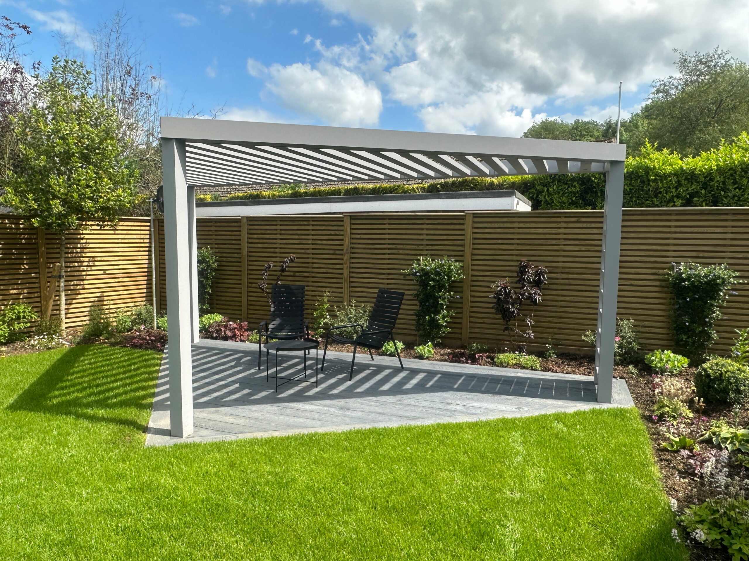 A modern garden pergola with two chairs sits on a lush green lawn, surrounded by landscaped plants and wooden fencing