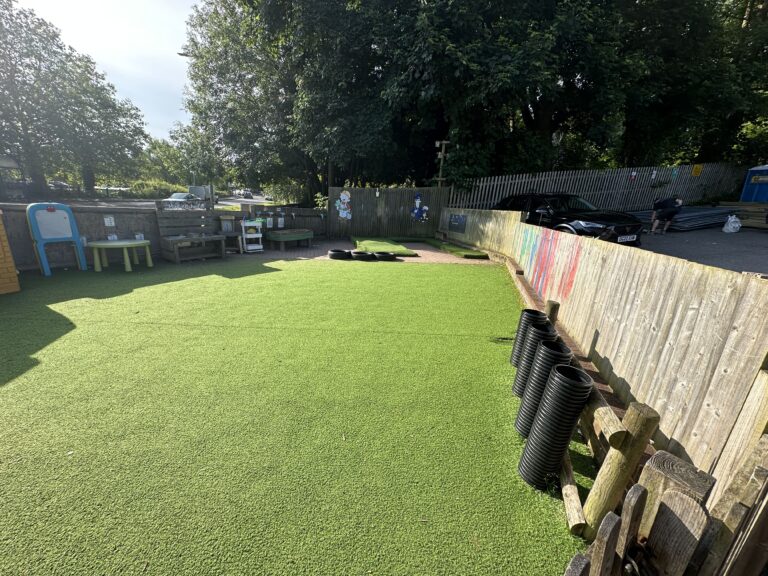 A vibrant green play area features artificial turf, wooden structures, and a fence, with sunlight filtering through trees