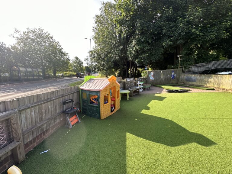 A playground scene featuring a colorful playhouse, green artificial turf, benches, and nearby trees, with a road in the background