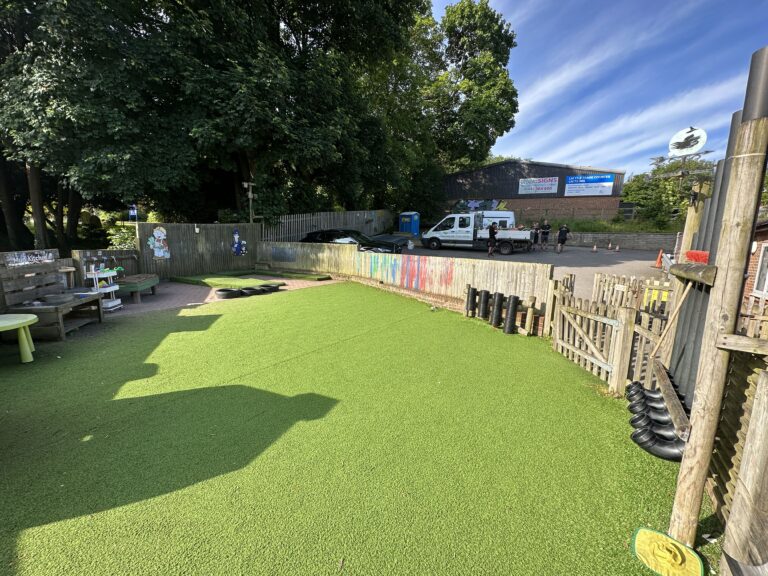 Bright playground with artificial grass, wooden seating, and a fenced area. Trees provide shade, while a building and vehicles are in the background