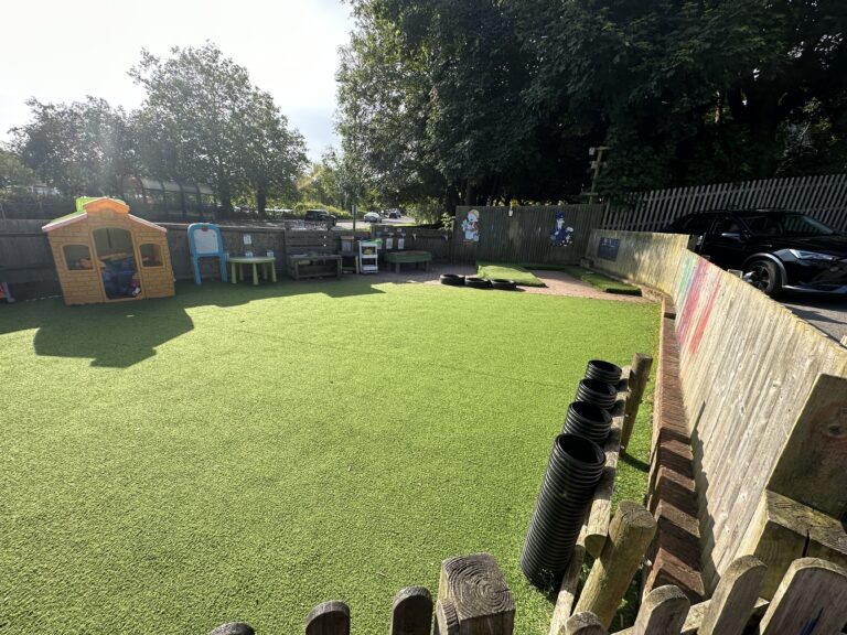 A vibrant play area featuring synthetic grass, a playhouse, seating, and toys, surrounded by a wooden fence