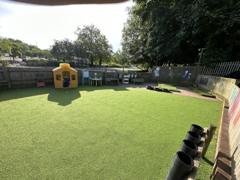 A bright, artificial turf play area with a small playhouse, tables, and colorful decorations on the fence