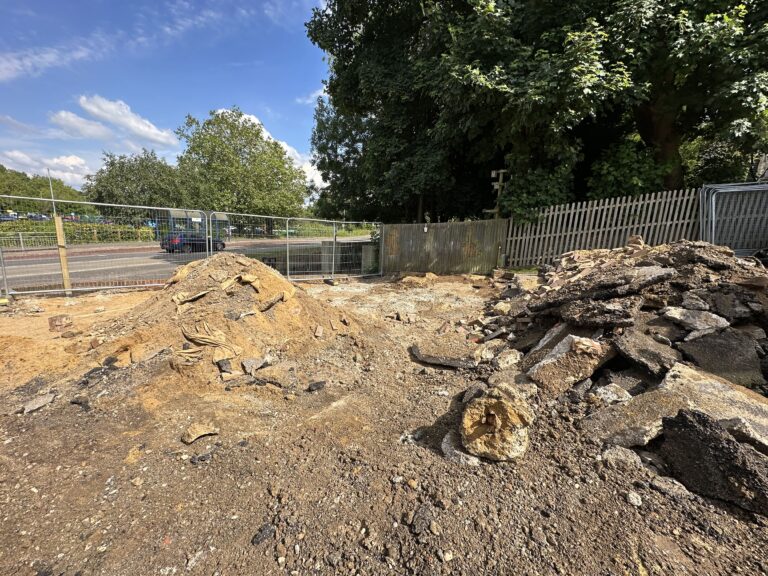 A construction site displays piles of dirt and debris, with a background of trees and a nearby road