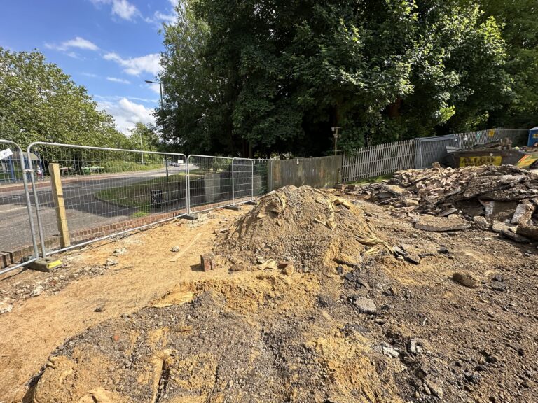 A construction site with a mound of dirt and debris, surrounded by fencing and trees under a blue sky