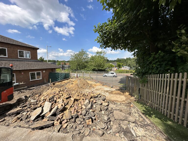 A construction site displaying a pile of rubble beside a brick building, with blue skies and greenery surrounding it