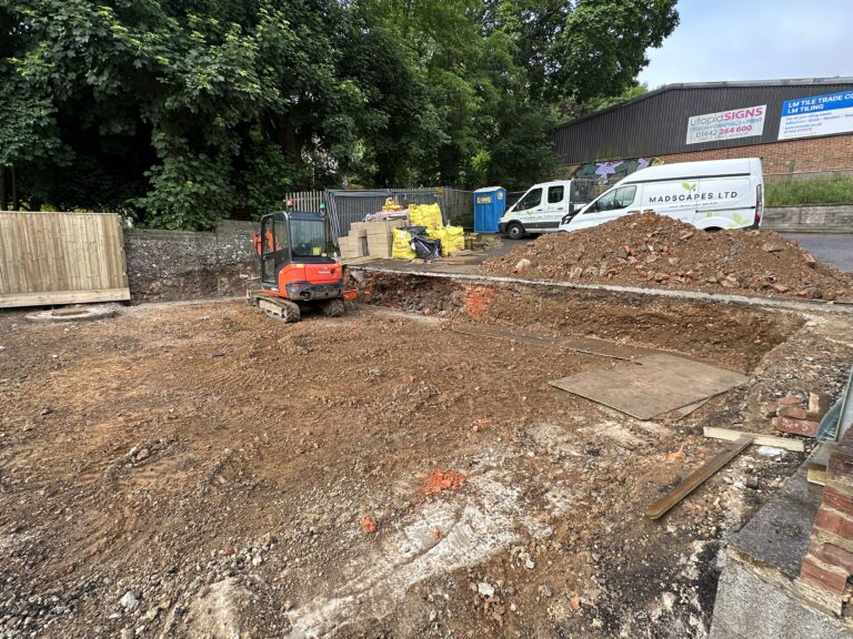 A construction site with a small excavator, dirt mound, and stacked materials. Trees and vehicles are visible in the background