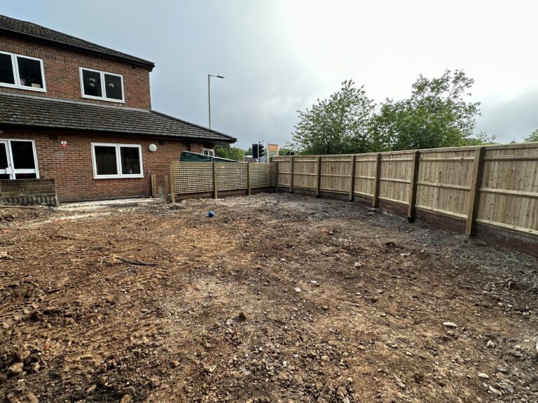 Empty, cleared outdoor area surrounded by new wooden fencing, next to a brick building under overcast skies
