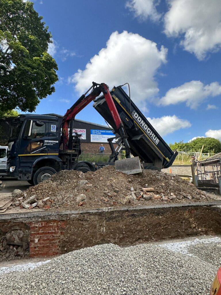 A construction site features a truck unloading materials, with cloudy blue skies and tree foliage in the background