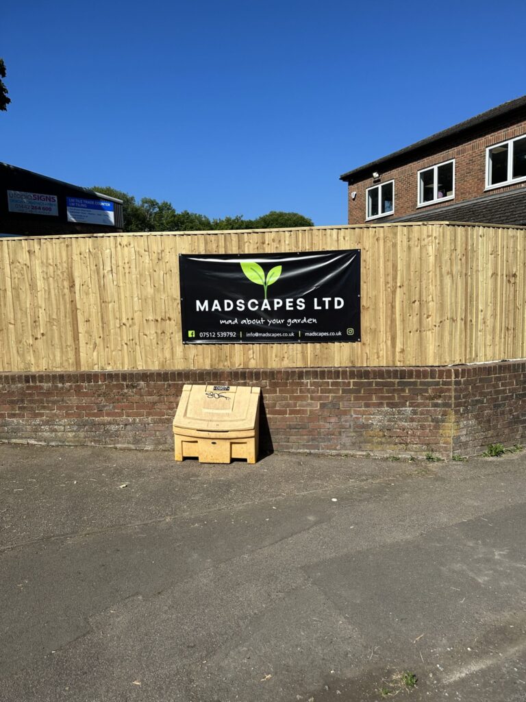 A wooden fence displays a large sign for "MADSCAPES LTD," promoting gardening services, with a clear blue sky overhead