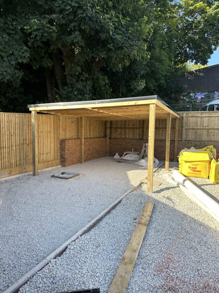 An unfinished outdoor structure with wooden framing, gravel flooring, and surrounding trees. Construction materials are nearby