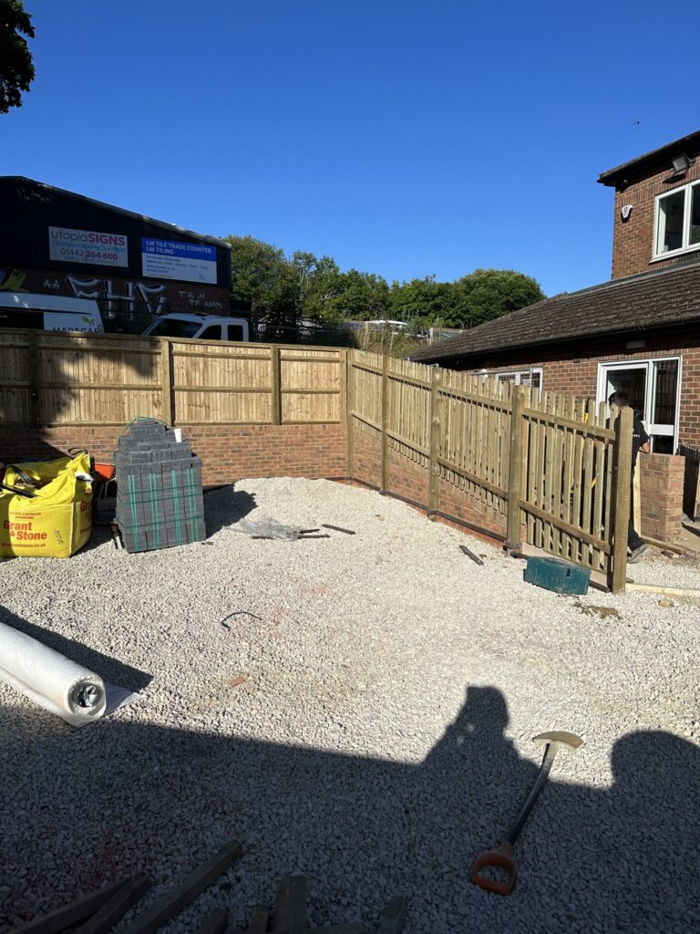 A construction site with gravel flooring, a wooden fence, and tools scattered around, under a clear blue sky