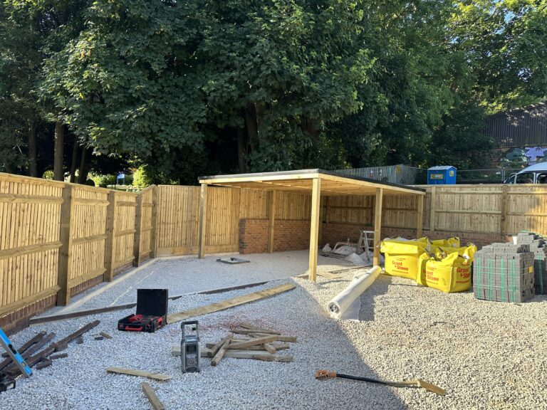 A construction site with a wooden frame structure, gravel flooring, tools, and building materials surrounded by wooden fencing and trees