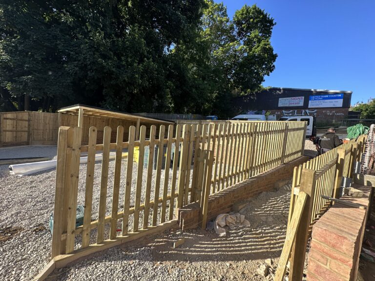 The image shows a newly constructed wooden fence alongside a gravel area, with trees in the background and some structures nearby