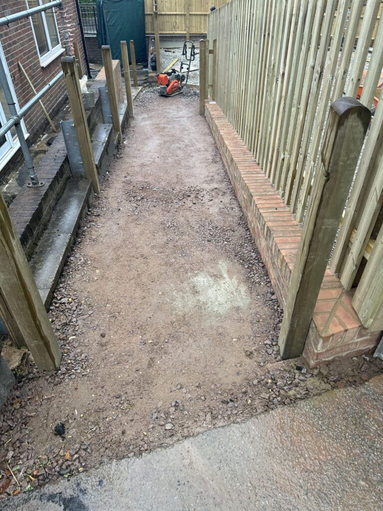 A narrow, unfinished pathway with gravel and wooden posts, flanked by fencing and construction equipment in the background
