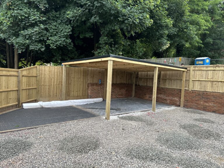 A partially constructed carport in a fenced area, with gravel flooring and trees in the background