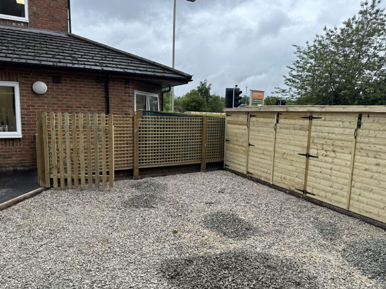 A gravel area features wooden fences and storage units beside a brick building under a cloudy sky