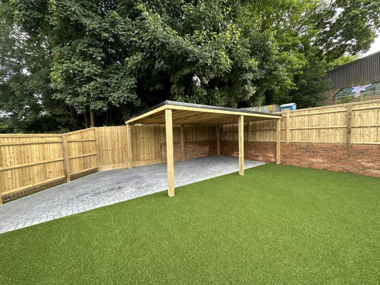 A well-maintained garden area features artificial grass, a brick wall, and a wooden-covered seating space. Lush trees surround the space