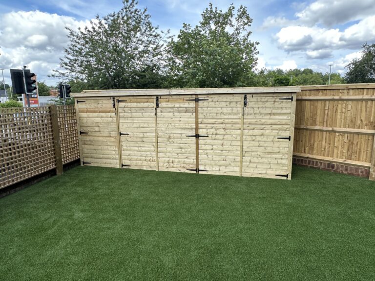 The image showcases a wooden storage shed with double doors, set on artificial grass, surrounded by a wooden fence