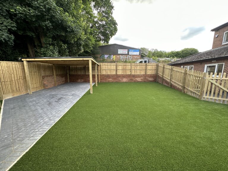 A spacious backyard features lush artificial grass, a paved area, and a wooden shelter against a fenced background