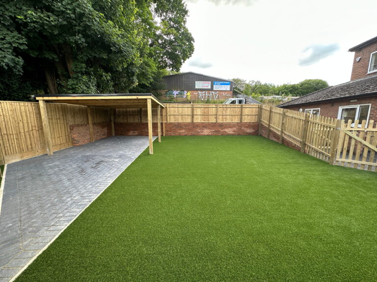 Spacious backyard featuring lush artificial grass, a brick wall, wooden fence, and a paved carport. Ideal for outdoor activities