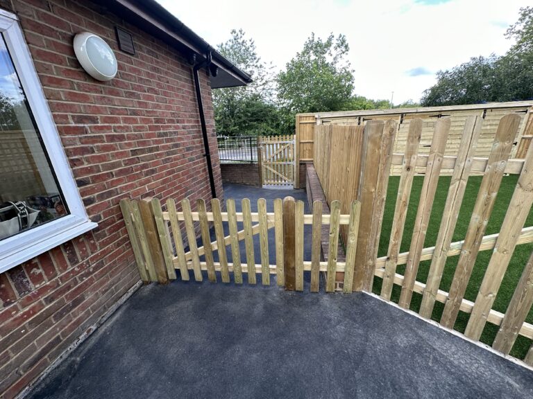 A wooden gate opens to a pathway beside a brick wall, leading to a fenced area with artificial grass