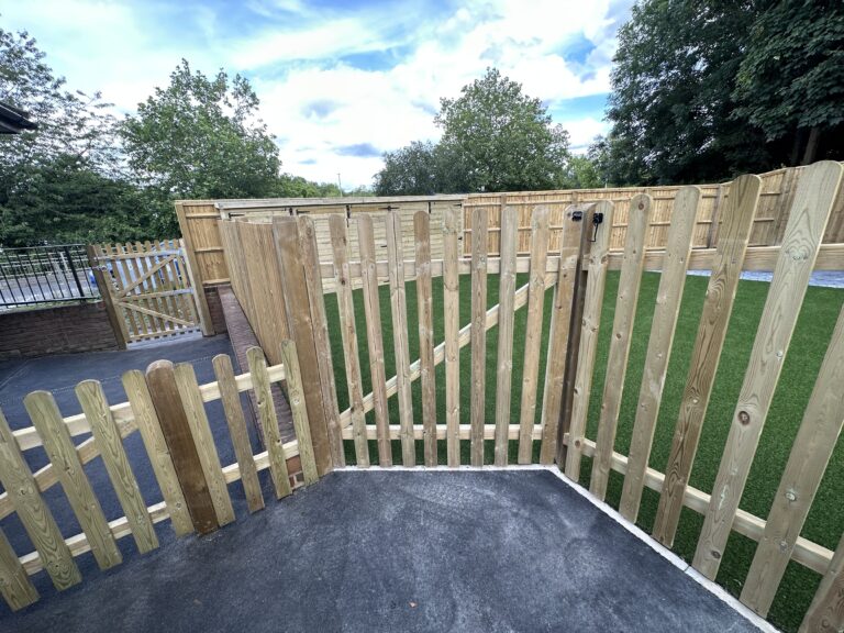 A wooden fenced area with a gate, lush green grass, and trees under a partly cloudy sky