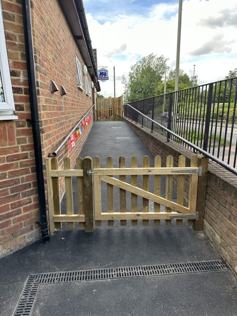 A wooden gate opens onto a paved pathway beside a brick building, bordered by a metal railing and greenery
