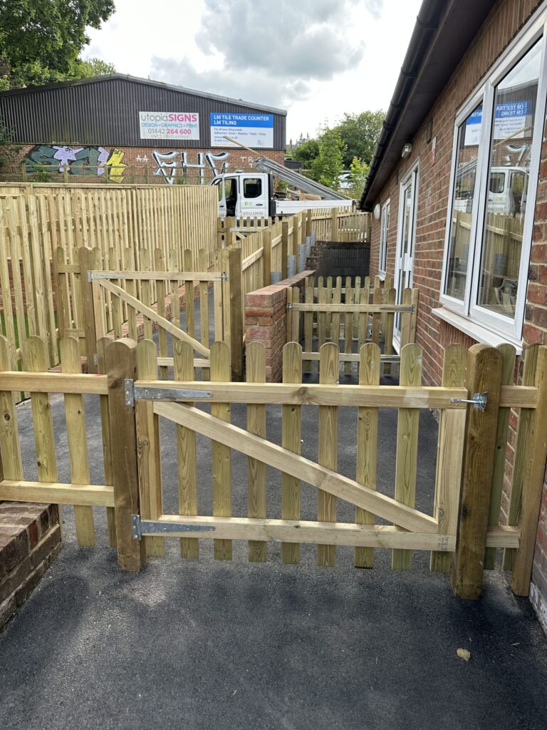 A wooden fence and gate structure beside a brick building, with a truck and colorful mural in the background