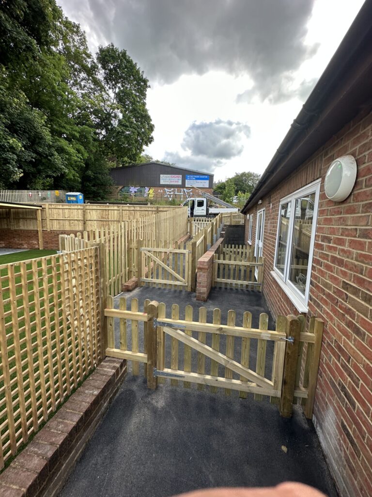 A wooden fenced area beside a brick structure, with pathways, trees, and partially cloudy skies in the background