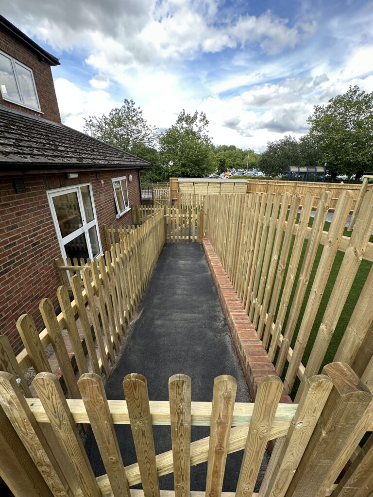 A wooden fence surrounds a paved walkway leading to a building, with trees and a partly cloudy sky in the background