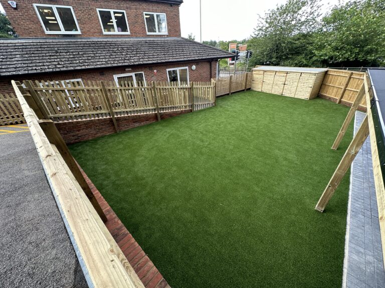 A spacious fenced outdoor area with artificial grass, adjacent to a brick building, featuring wooden barriers and storage sheds