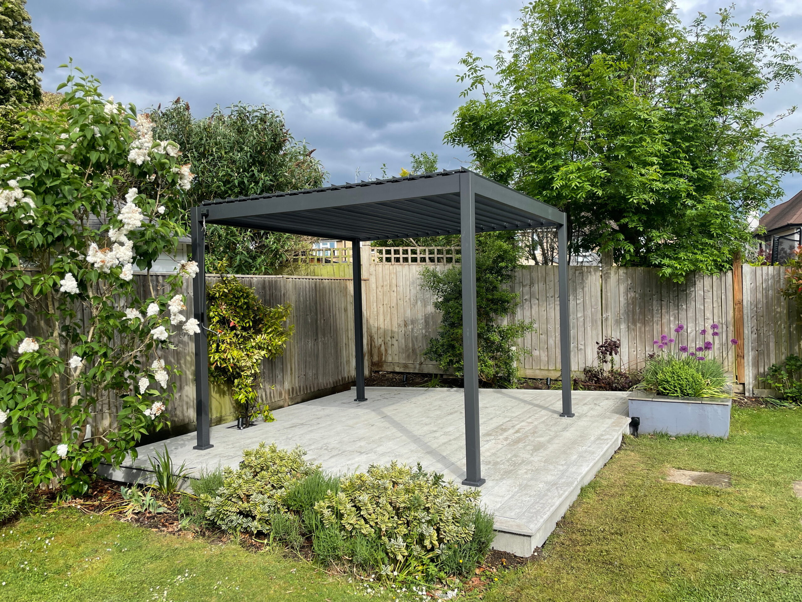 Modern garden pergola surrounded by vibrant flowers and greenery, set against a cloudy sky and wooden fence