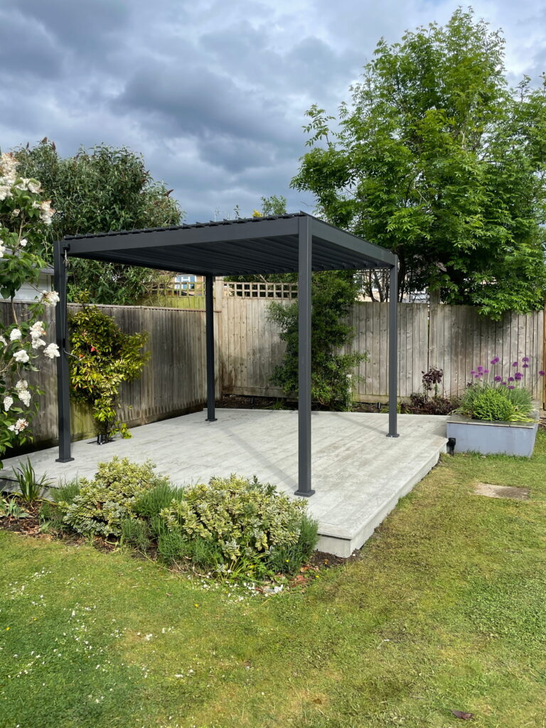 Modern garden gazebo on a concrete patio, surrounded by lush greenery and colorful flowers under a cloudy sky