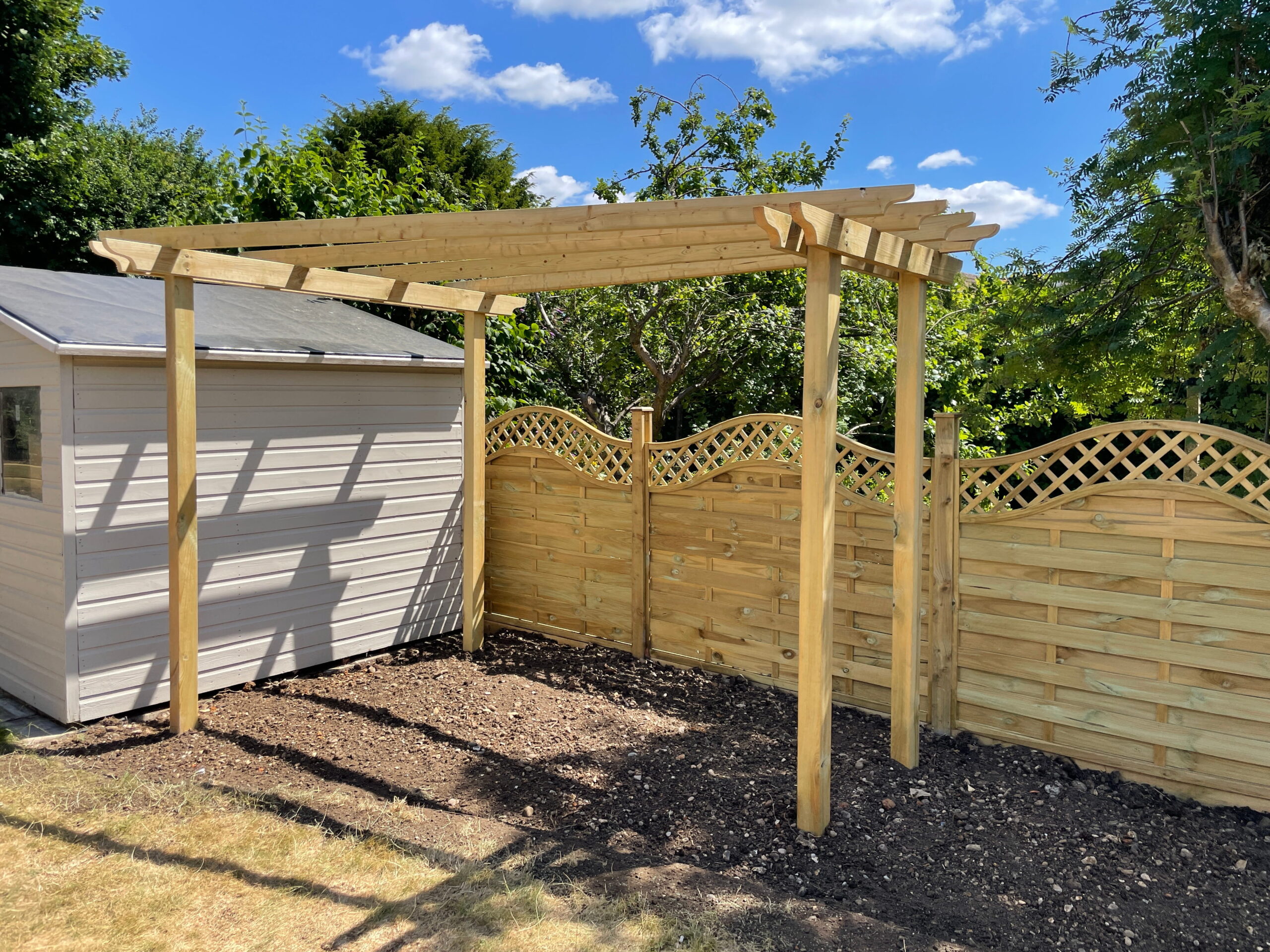 Wooden pergola beside a garden shed and decorative fencing, set against a bright blue sky. Ideal for outdoor projects