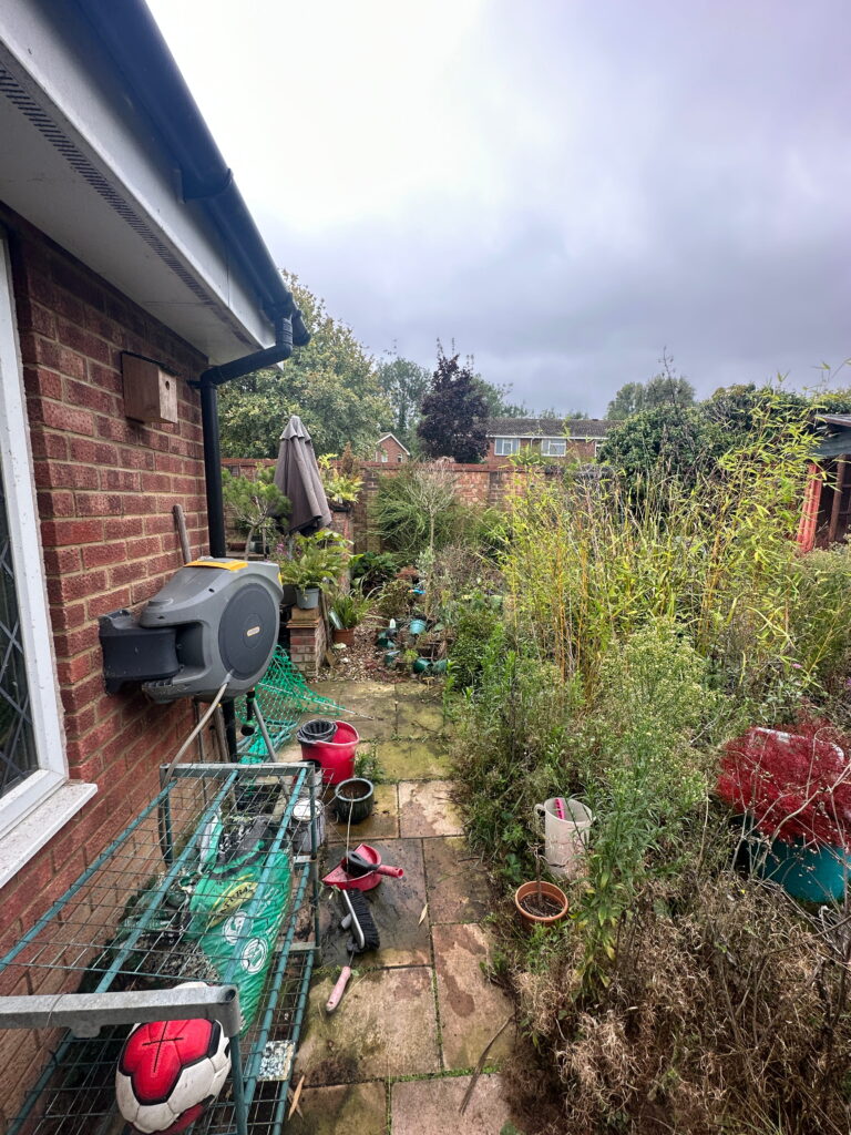 A cluttered garden area featuring overgrown plants, garden tools, and various pots, under a cloudy sky