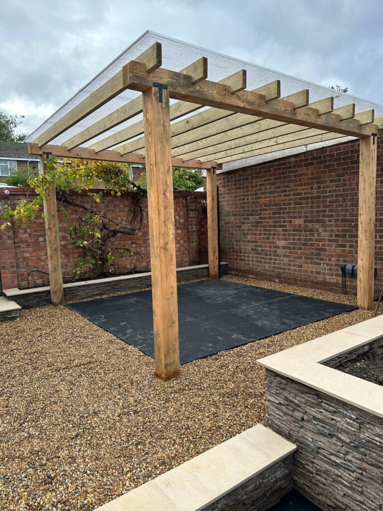 A wooden pergola with a covered roof stands over a gravel surface in a newly landscaped garden area